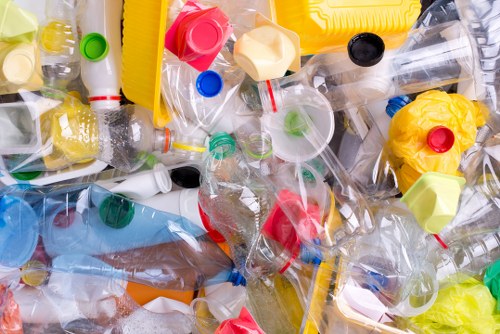 A close-up view of a pile of discarded plastic bottles and containers, including some with caps of various colors such as green, red, blue, yellow, and black. The bottles are made of clear PET plastic and are crumpled or flattened, with some partially crushed, indicating they have been used and discarded. Among the bottles, there are also plastic lids, caps, and a few pieces of plastic wrap or packaging material, all scattered randomly across a surface, likely on a driveway or waste collection area. The background shows more plastic waste, with some parts overlapping, creating a colorful and textured mixture of materials. This scene represents typical household or commercial plastic waste awaiting collection, consistent with rubbish removal services in Teddington, where Commercial Waste Removal Teddington operates, offering waste disposal solutions for plastics and other refuse materials.