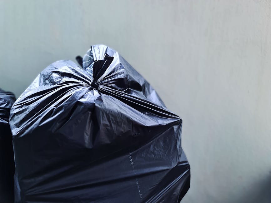 Close-up of several black rubbish bags made of glossy plastic material, tightly knotted at the top, positioned in the foreground against a neutral light grey wall. The bags are stacked closely togethe