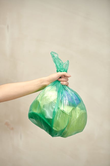 Close-up of several black rubbish bags made of glossy plastic material, tightly knotted at the top, positioned in the foreground against a neutral light grey wall. The bags are stacked closely togethe