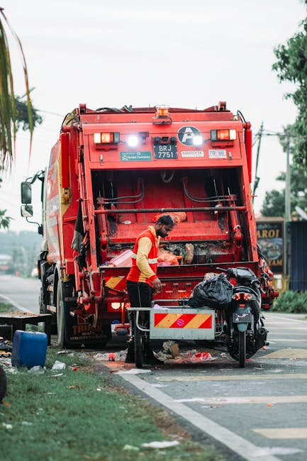 A person is disposing of crumpled brown paper bags into a grey bin that is designated for waste collection, positioned on a wooden surface. The person's arm, wearing a short-sleeved white top, is visi